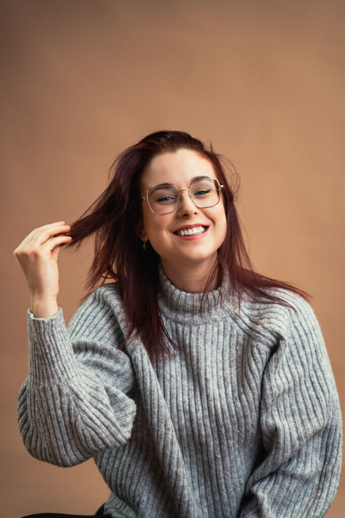 Junge Frau mit Brille und lockigem, rotem Haar in einem grauen Pullover lächelt und spielt mit ihren Haaren vor einem bei...