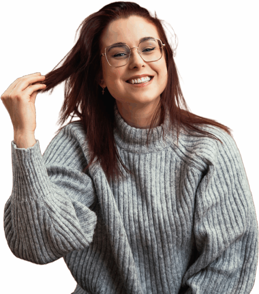Junge Frau mit roten Haaren und Brille, die lächelt und spielerisch an ihren Haaren zieht, trägt einen grauen Pullover.